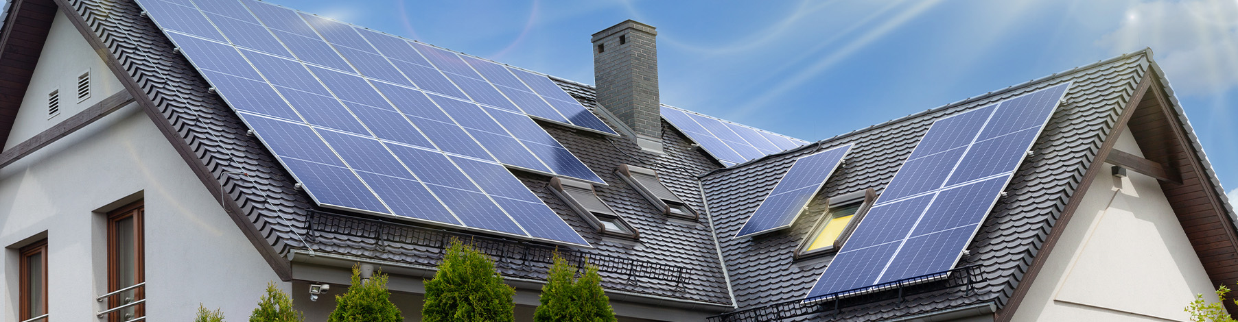Eine Panoramabildaufnahme eines Einfamilienhauses. Das dunkle, geneigte Dach ist fast vollständig mit blauen Solarmodulen bedeckt, die in verschiedenen Sektionen angeordnet sind. Der Himmel ist blau mit einigen Wolken und die Sonne scheint auf die Module.