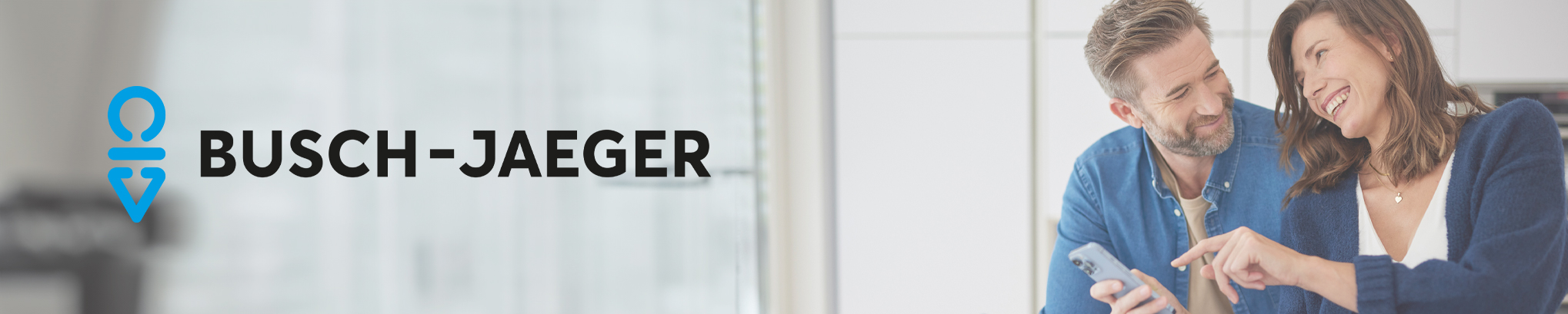 Im Hintergrund ist unscharf auf der rechten Seite ein lächelndes Paar in einer Küche zu sehen, das ein Smartphone bedient. Im Vordergrund ist das Busch-Jaeger Logo links zu sehen.