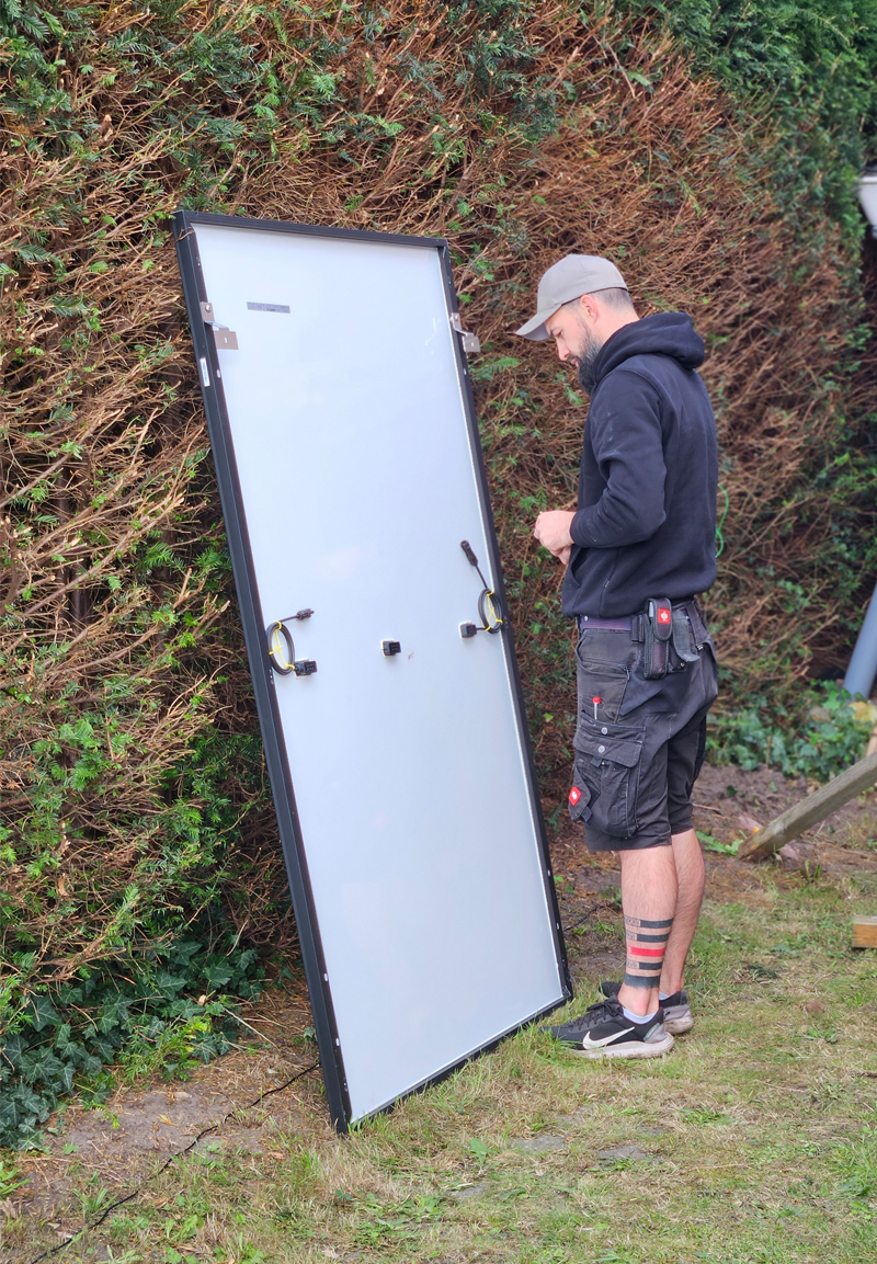 Ein Mann mit schwarzen Klamotten und schwarzem Basecap steht vor einem Modul, dessen Rückseite zu sehen ist und an einer Hecke lehnt