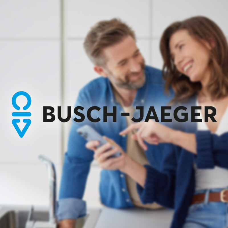 Im Hintergrund ist unscharf auf der rechten Seite ein lächelndes Paar in einer Küche zu sehen, das ein Smartphone bedient. Im Vordergrund ist das Busch-Jaeger Logo links zu sehen.