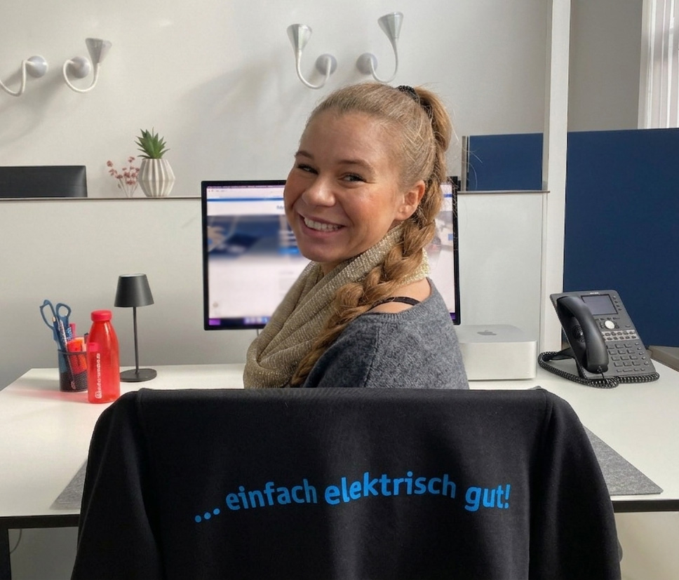 Eine lächelnde Mitarbeiterin an einem modern ausgestatteten Büro-Arbeitsplatz bei elektroland24. Im Vordergrund hängt ein schwarzes Kleidungsstück über dem Stuhl mit dem blauen Firmenslogan „... einfach elektrisch gut!“.