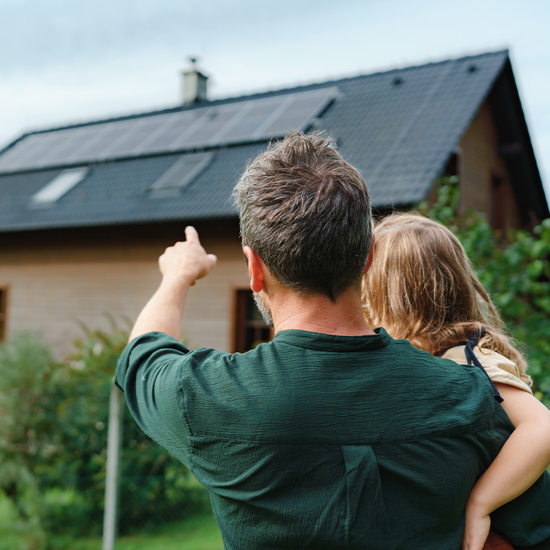 Ein Mann hält ein Kind auf dem Arm und zeigt auf die Photovoltaikanlage auf dem Hausdach – Symbolbild für die Erweiterung einer PV-Anlage für die Zukunft.