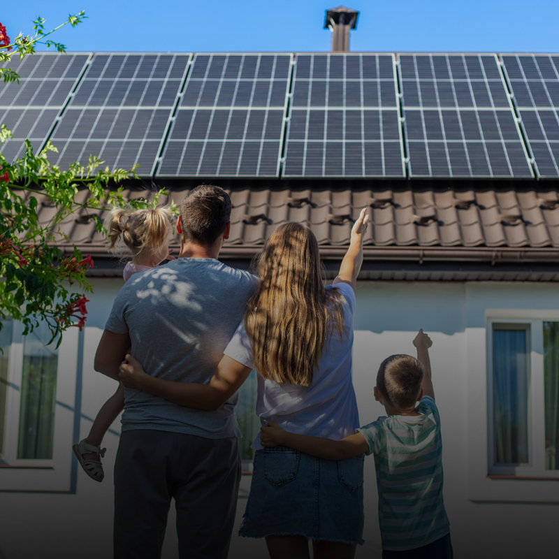 Eltern stehen mit ihren zwei Kindern vor einem Haus mit Solarpanelen und zeigen mit dem rechten Finger darauf