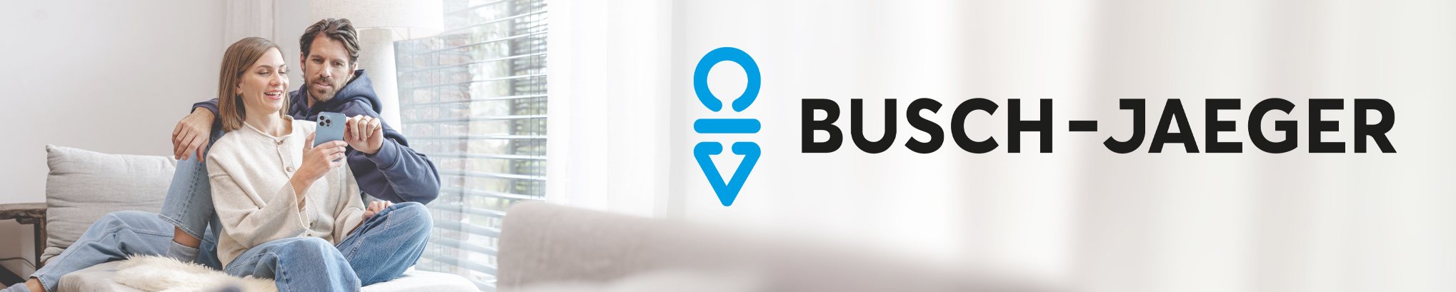 Ein Paar sitzt entspannt auf einem Sofa und bedient gemeinsam ein Smartphone. Rechts daneben steht das Busch-Jaeger Logo auf hellem, minimalistischem Hintergrund.