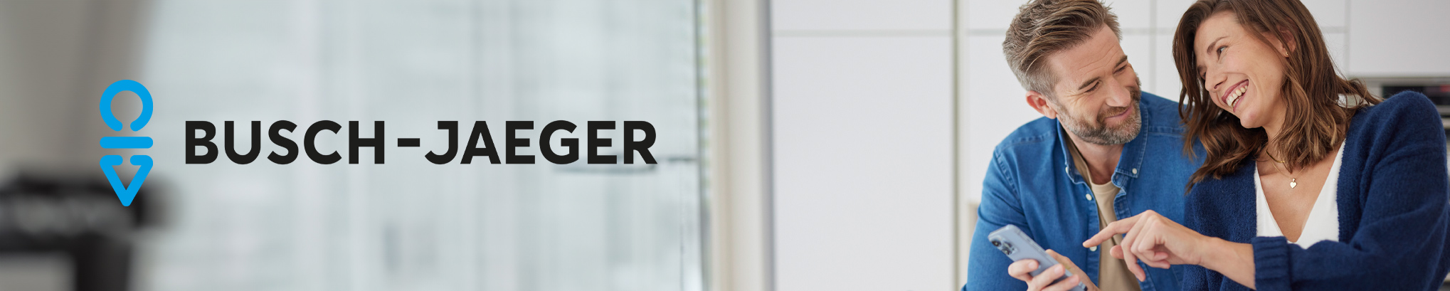 Im Hintergrund ist unscharf auf der rechten Seite ein lächelndes Paar in einer Küche zu sehen, das ein Smartphone bedient. Im Vordergrund ist das Busch-Jaeger Logo links zu sehen.