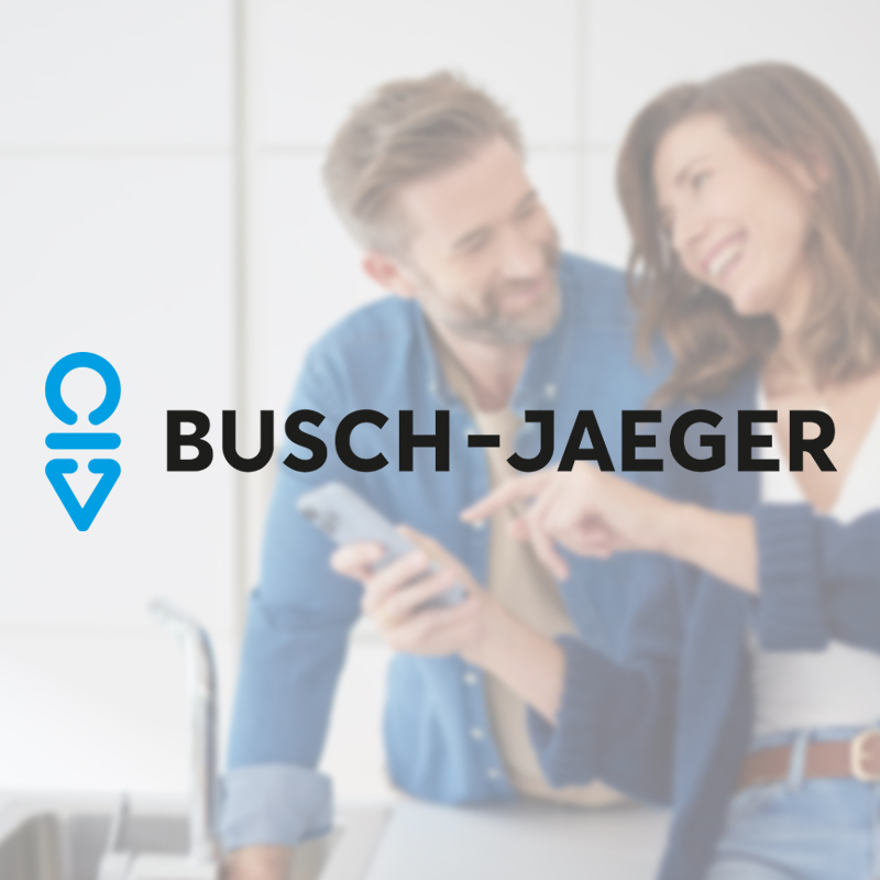 Im Hintergrund ist unscharf ein lächelndes Paar in einer Küche zu sehen, das ein Smartphone bedient. Im Vordergrund ist das Busch-Jaeger Logo mittig zu sehen.